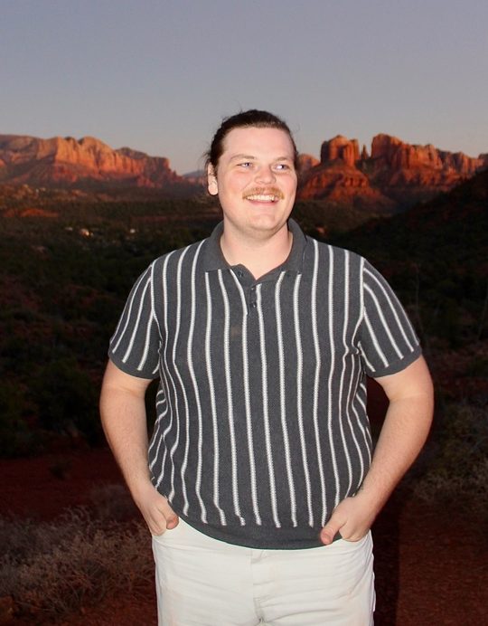 Jackson grinning at dusk with his hands in his pockets, red rock cliffs behind him